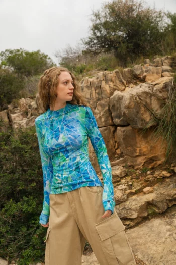 Kobieta o falujących jasnobrązowych włosach stoi na zewnątrz w pobliżu skalistego terenu, ubrana w niebiesko-zieloną wzorzystą bluzkę z długimi rękawami i beżowe spodnie cargo. Spogląda w bok, otoczona naturalną zielenią i skałami.
