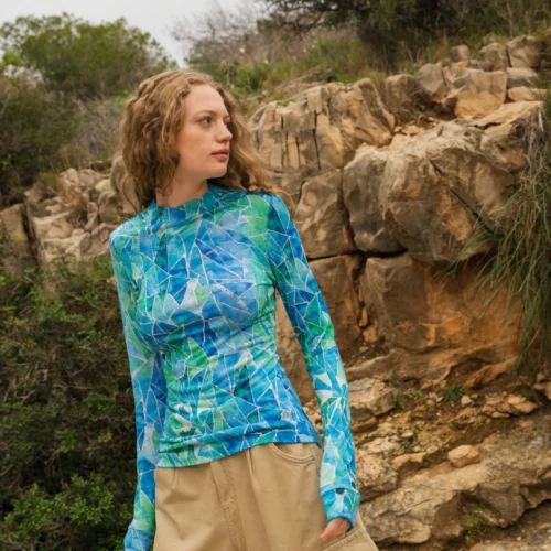 Kobieta o falujących jasnobrązowych włosach stoi na zewnątrz w pobliżu skalistego terenu, ubrana w niebiesko-zieloną wzorzystą bluzkę z długimi rękawami i beżowe spodnie cargo. Spogląda w bok, otoczona naturalną zielenią i skałami.
