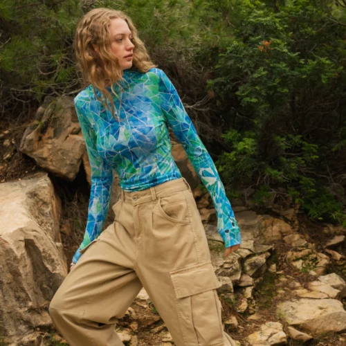 Osoba o falujących blond włosach ubrana w niebieską wzorzystą bluzkę z długim rękawem i luźne spodnie cargo stoi na skalistej ścieżce otoczonej zielenią.