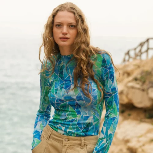 Kobieta z długimi falującymi włosami ma na sobie niebiesko-zieloną bluzkę z długim rękawem w geometryczne wzory i beżowe spodnie. Stoi na zewnątrz w pobliżu skalistego wybrzeża, z oceanem i drewnianym płotem widocznym w tle.