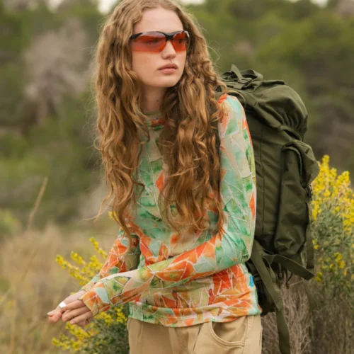 Osoba z długimi falowanymi włosami w pomarańczowych okularach przeciwsłonecznych, kolorowej koszuli z długim rękawem i beżowych spodniach stoi na zewnątrz z zielonym plecakiem. W tle widoczne są żółte kwiaty i zieleń.