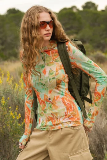 Osoba z długimi falującymi włosami i pomarańczowymi okularami przeciwsłonecznymi ma na sobie kolorową koszulę z długim rękawem w geometryczne wzory i beżowe spodnie cargo, niosąc zielony plecak, stojąc na zewnątrz w trawiastym, zalesionym obszarze.