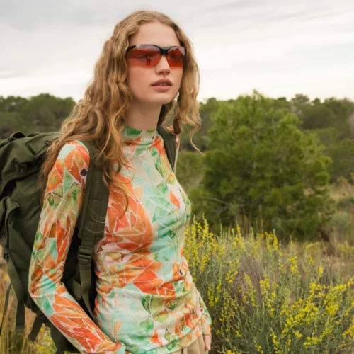 Kobieta z falistymi blond włosami w pomarańczowych okularach przeciwsłonecznych, kolorowej koszuli z długim rękawem w geometryczne wzory, beżowych spodniach i zielonym plecaku stoi na zewnątrz w naturalnym otoczeniu z zielenią i zachmurzonym niebem.
