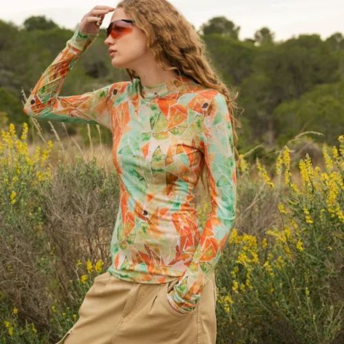 Osoba z długimi kręconymi włosami nosi pomarańczowe okulary przeciwsłoneczne, kolorową koszulę z długim rękawem z zielonymi i pomarańczowymi wzorami oraz beżowe spodnie, stojąc przed wysokimi zielonymi krzewami i żółtymi polnymi kwiatami na zewnątrz.