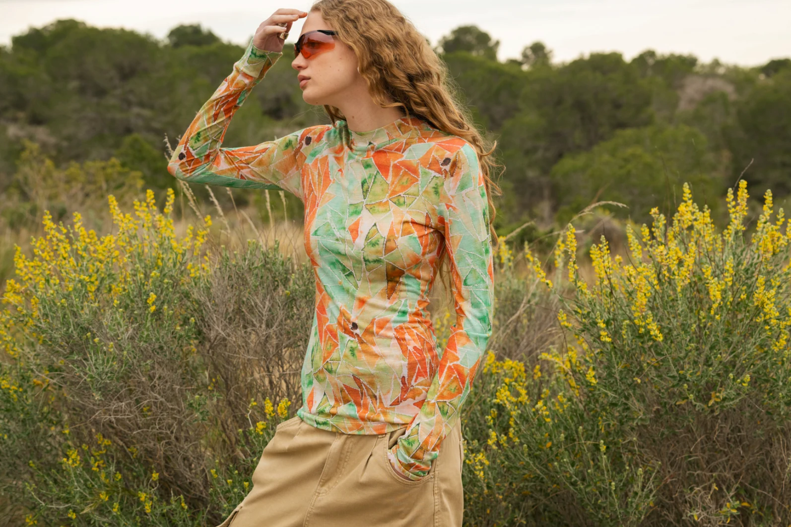 Osoba z długimi kręconymi włosami nosi pomarańczowe okulary przeciwsłoneczne, kolorową koszulę z długim rękawem z zielonymi i pomarańczowymi wzorami oraz beżowe spodnie, stojąc przed wysokimi zielonymi krzewami i żółtymi polnymi kwiatami na zewnątrz.