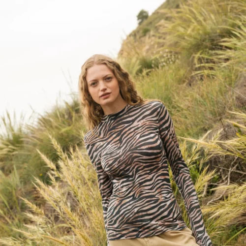 Kobieta z falującymi włosami stoi na zewnątrz na trawiastym wzgórzu, ubrana w bluzkę z długim rękawem w zebrę i beżowe spodnie. W tle widać wysoką trawę i pochyłe wzgórze pod zachmurzonym niebem.