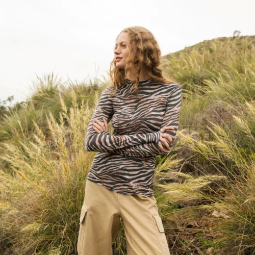 Osoba o falowanych blond włosach ubrana w bluzkę z długim rękawem w zebrę i beżowe spodnie stoi ze skrzyżowanymi ramionami przed wysoką trawą i zboczem wzgórza w pochmurny dzień.