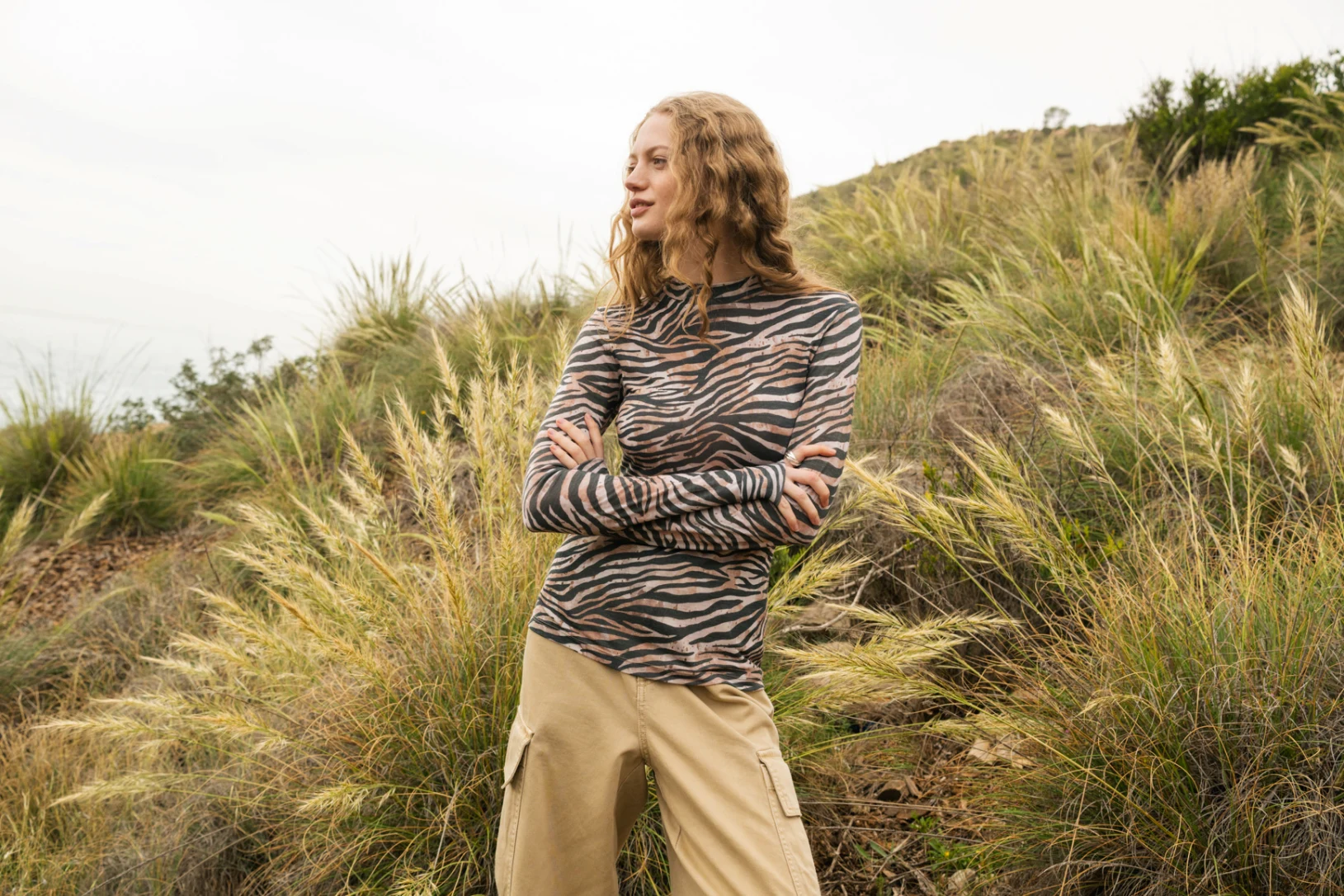 Osoba o falowanych blond włosach ubrana w bluzkę z długim rękawem w zebrę i beżowe spodnie stoi ze skrzyżowanymi ramionami przed wysoką trawą i zboczem wzgórza w pochmurny dzień.