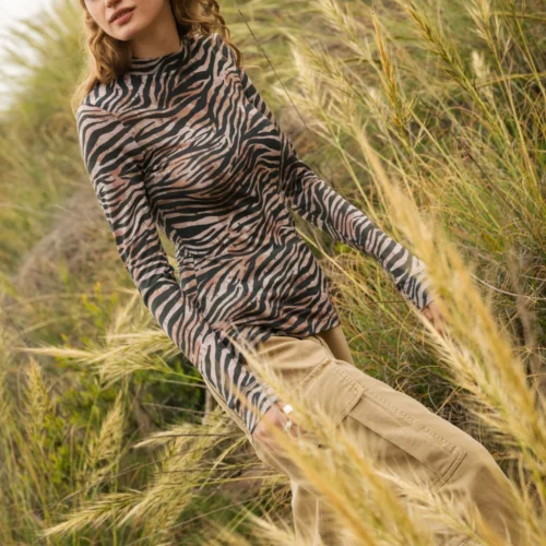 Kobieta z falującymi włosami ubrana w bluzkę z długim rękawem w zebrę i beżowe spodnie cargo stoi wśród wysokich traw, patrząc w bok ze spokojnym wyrazem twarzy.