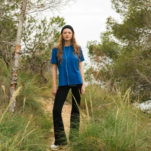 Kobieta z długimi włosami, ubrana w niebieską koszulę, czarne spodnie, białe buty i czarną czapkę, stoi na wąskiej polnej ścieżce otoczonej trawą i drzewami.