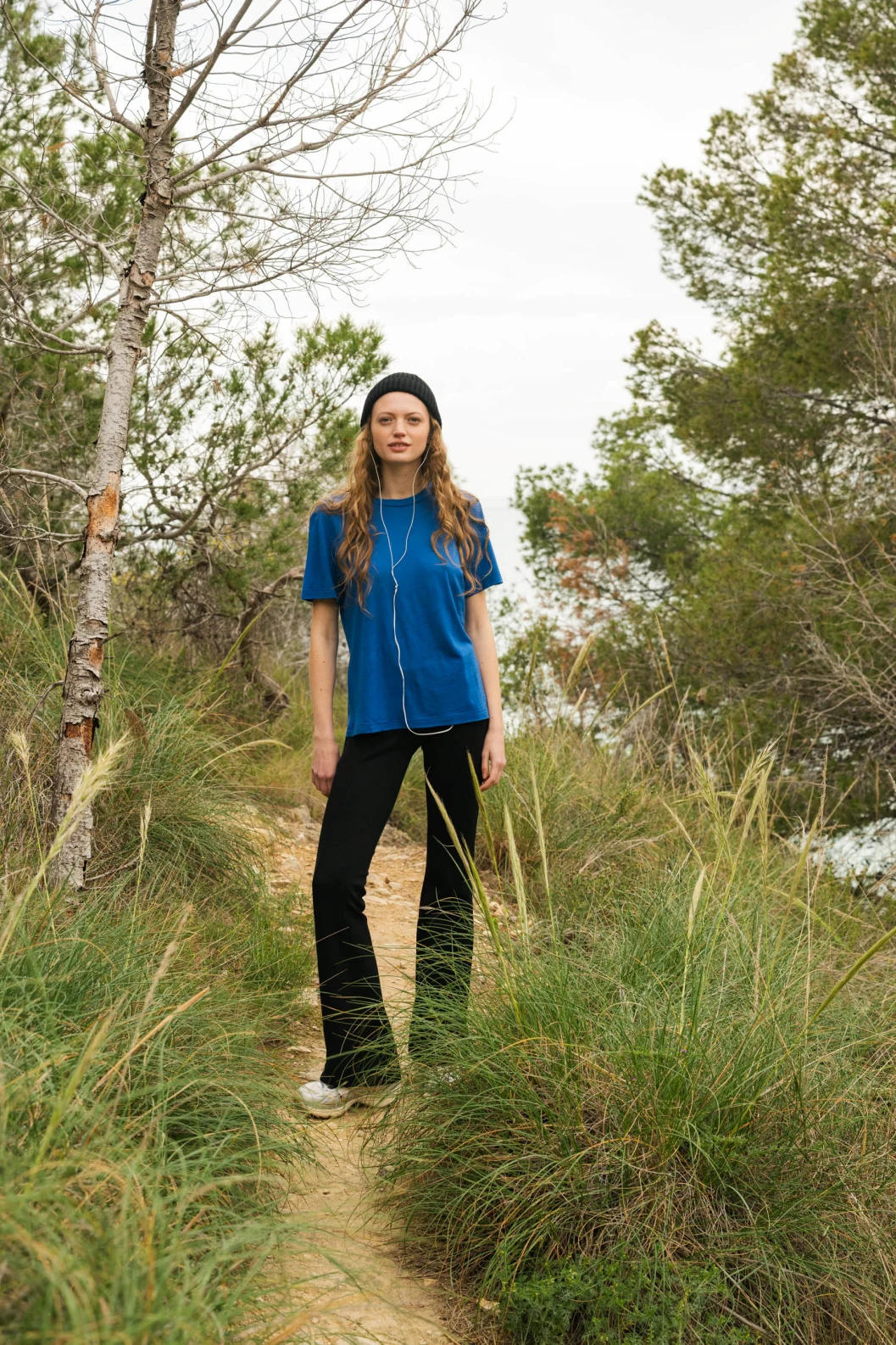 Kobieta z długimi włosami, ubrana w niebieską koszulę, czarne spodnie, białe buty i czarną czapkę, stoi na wąskiej polnej ścieżce otoczonej trawą i drzewami.