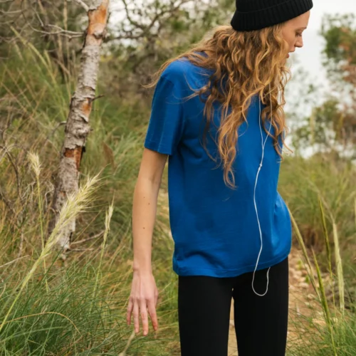 Osoba z długimi falującymi włosami, ubrana w niebieską koszulkę, czarne spodnie i czarną czapkę, stoi na zewnątrz wśród wysokiej trawy i drzew, patrząc w dół ze słuchawkami zawieszonymi na szyi.