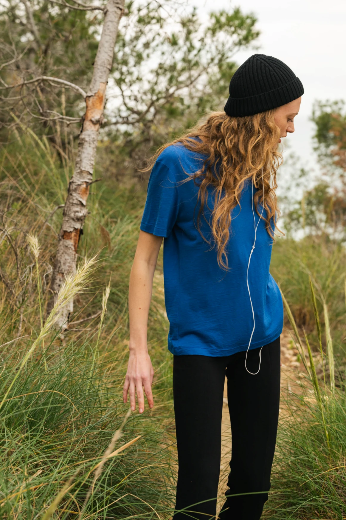 Osoba z długimi falującymi włosami, ubrana w niebieską koszulkę, czarne spodnie i czarną czapkę, stoi na zewnątrz wśród wysokiej trawy i drzew, patrząc w dół ze słuchawkami zawieszonymi na szyi.