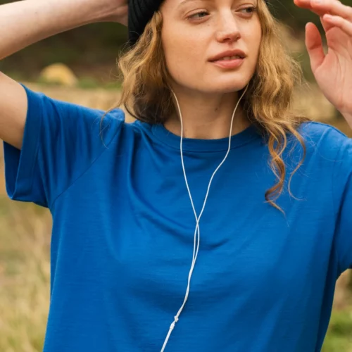 Kobieta w niebieskiej koszulce i czarnej czapce stoi na zewnątrz, słuchając muzyki przez białe słuchawki. Ma jedną rękę za głową i patrzy w bok, z zrelaksowanym wyrazem twarzy.