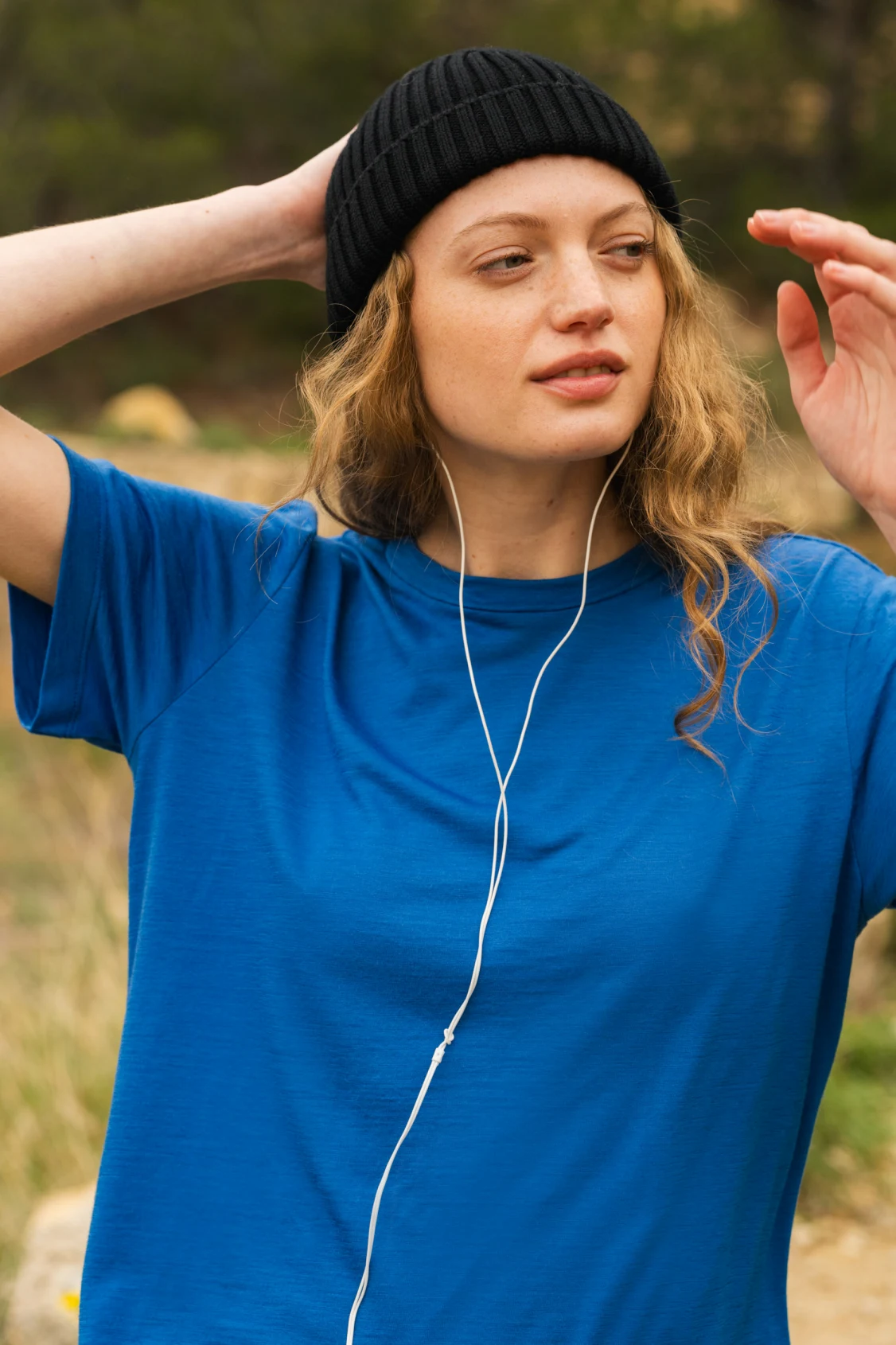 Kobieta w niebieskiej koszulce i czarnej czapce stoi na zewnątrz, słuchając muzyki przez białe słuchawki. Ma jedną rękę za głową i patrzy w bok, z zrelaksowanym wyrazem twarzy.