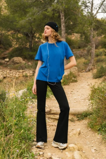 Osoba ubrana w niebieską koszulkę, czarne spodnie, białe trampki i czarną czapkę stoi na kamienistej ścieżce w naturalnym otoczeniu z drzewami i trawą, słuchając słuchawek i patrząc w bok.