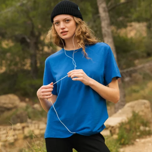 Osoba ubrana w niebieską koszulkę i czarną czapkę stoi na zewnątrz w zalesionym obszarze, trzymając białe słuchawki. W tle widać drzewa i polną ścieżkę.