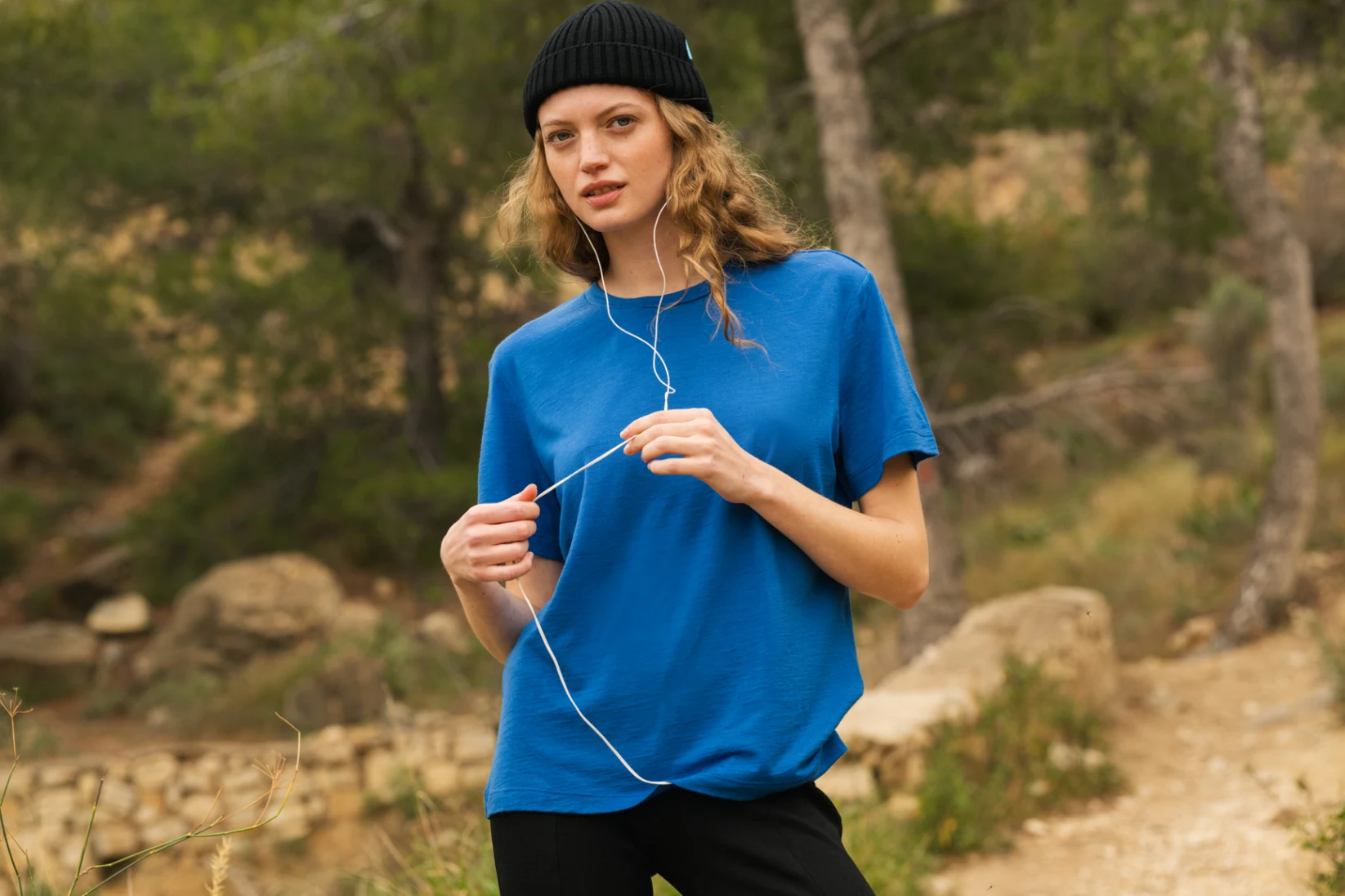 Osoba ubrana w niebieską koszulkę i czarną czapkę stoi na zewnątrz w zalesionym obszarze, trzymając białe słuchawki. W tle widać drzewa i polną ścieżkę.