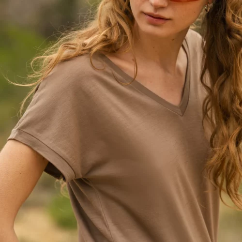 Kobieta z długimi falowanymi włosami ma na sobie oliwkowo-brązową koszulkę z dekoltem w szpic, pasującą opaskę na głowę i okulary przeciwsłoneczne w pomarańczowym odcieniu, stojąc na zewnątrz z zielenią w tle.