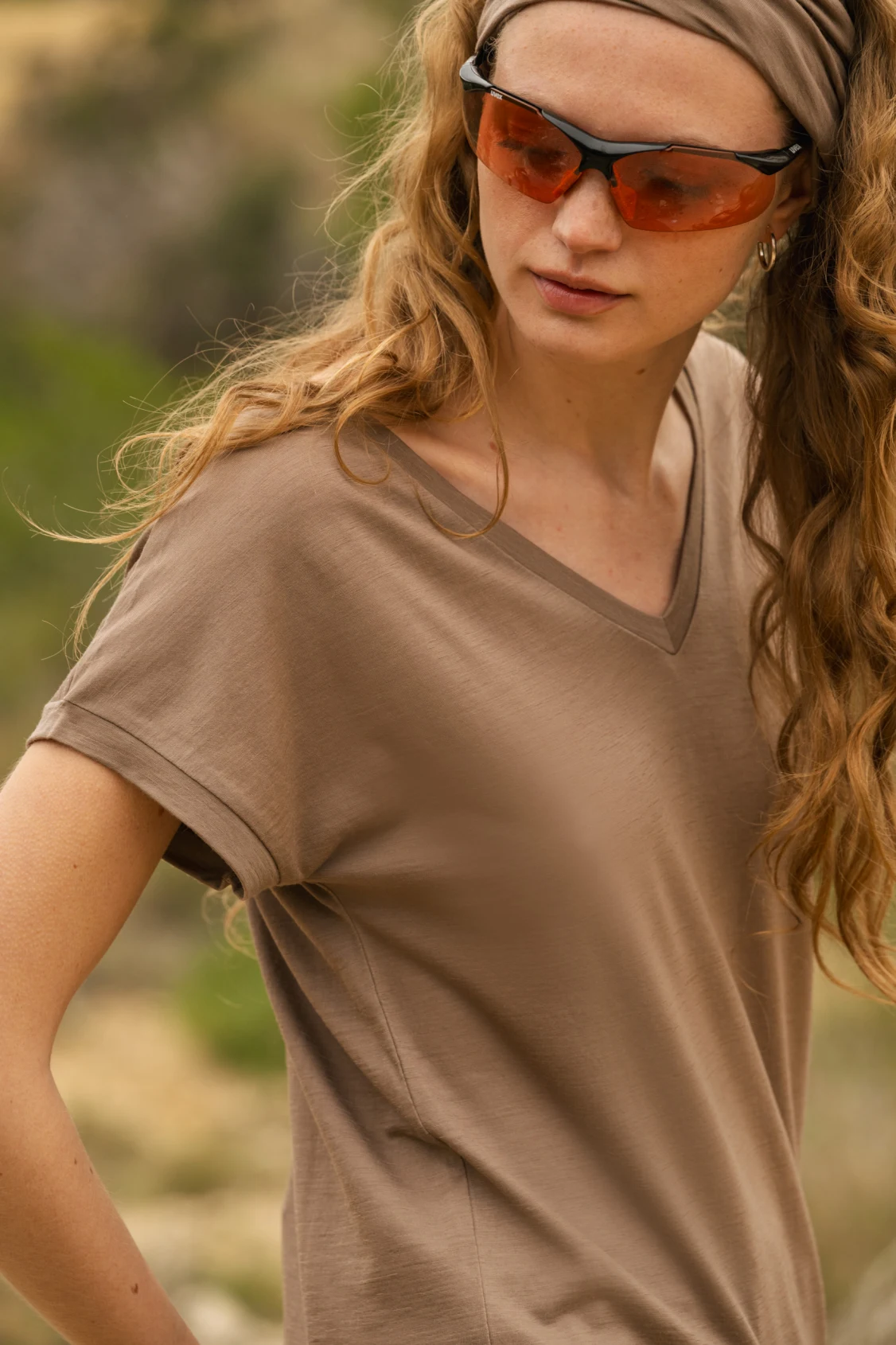 Kobieta z długimi falowanymi włosami ma na sobie oliwkowo-brązową koszulkę z dekoltem w szpic, pasującą opaskę na głowę i okulary przeciwsłoneczne w pomarańczowym odcieniu, stojąc na zewnątrz z zielenią w tle.