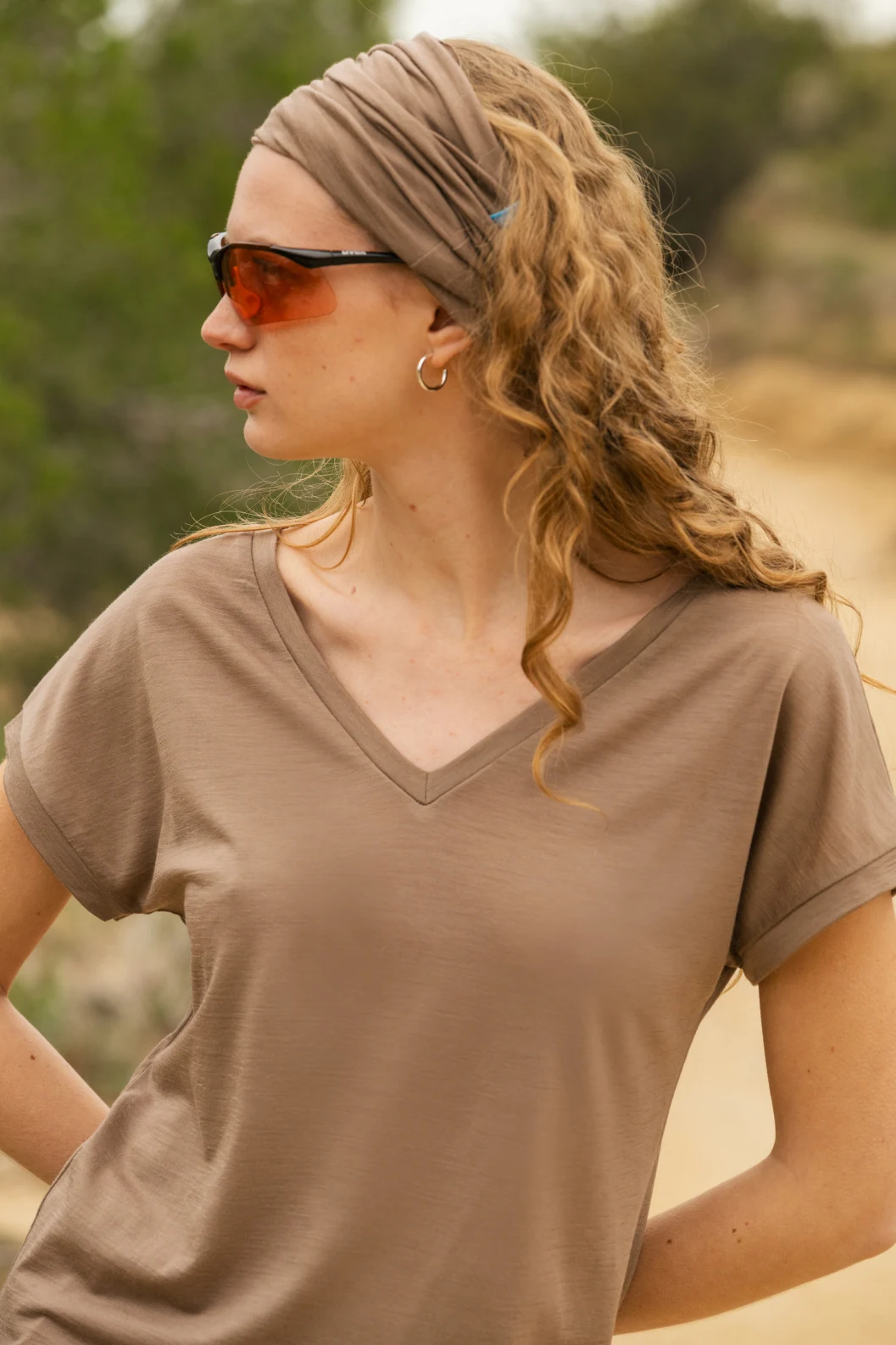 Kobieta z falującymi blond włosami ma na sobie jasnobrązową opaskę, brązowe okulary przeciwsłoneczne, kolczyki w kształcie obręczy i ciemnoszarą koszulę z dekoltem w szpic. Stoi na zewnątrz, patrząc w bok, z drzewami i piaszczystą ścieżką w tle.