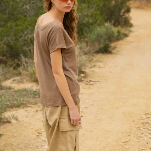 Kobieta w okularach przeciwsłonecznych, brązowej koszuli, beżowych spodniach cargo i chustce na głowie stoi na polnej ścieżce na zewnątrz, otoczona zielonymi krzewami i drzewami.