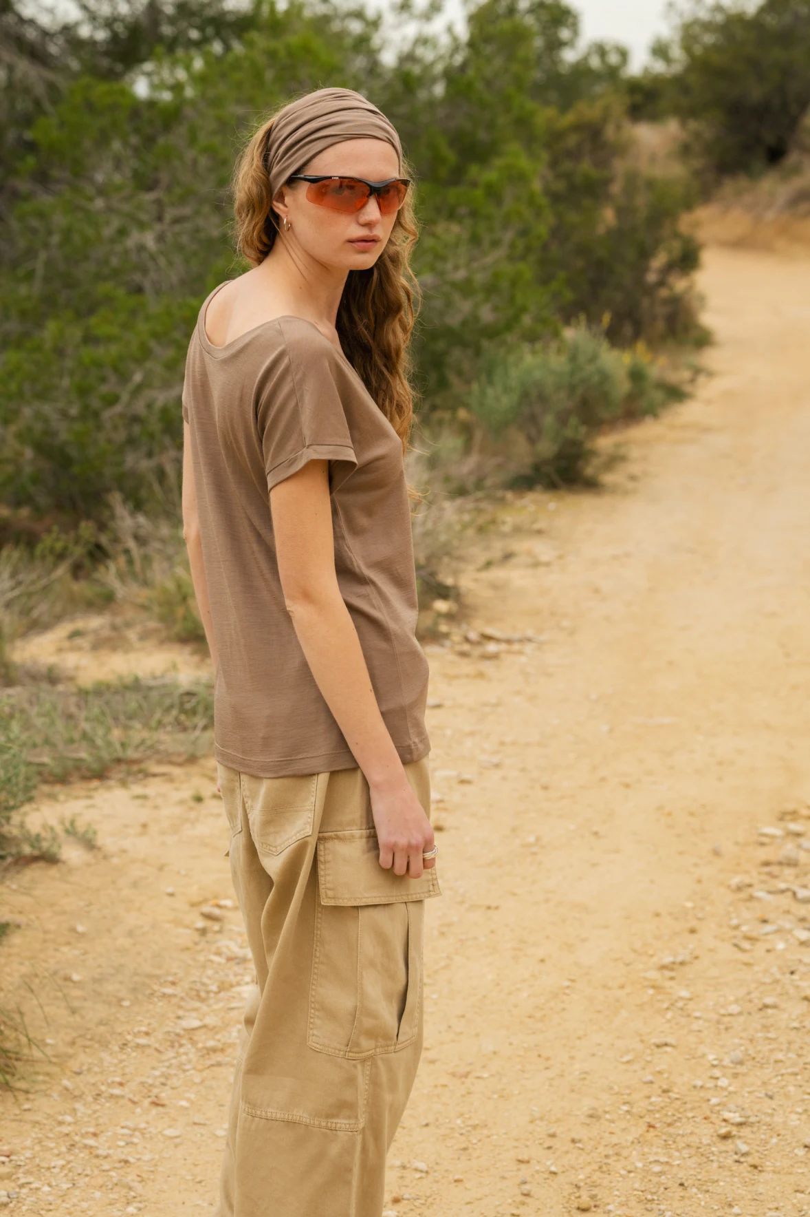 Kobieta w okularach przeciwsłonecznych, brązowej koszuli, beżowych spodniach cargo i chustce na głowie stoi na polnej ścieżce na zewnątrz, otoczona zielonymi krzewami i drzewami.