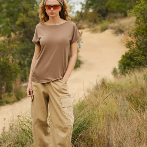 Osoba ubrana w szarą koszulkę, beżowe spodnie cargo, opaskę na głowę i pomarańczowe okulary przeciwsłoneczne stoi na zewnątrz na polnej ścieżce otoczonej wysoką trawą i zielonymi krzewami.