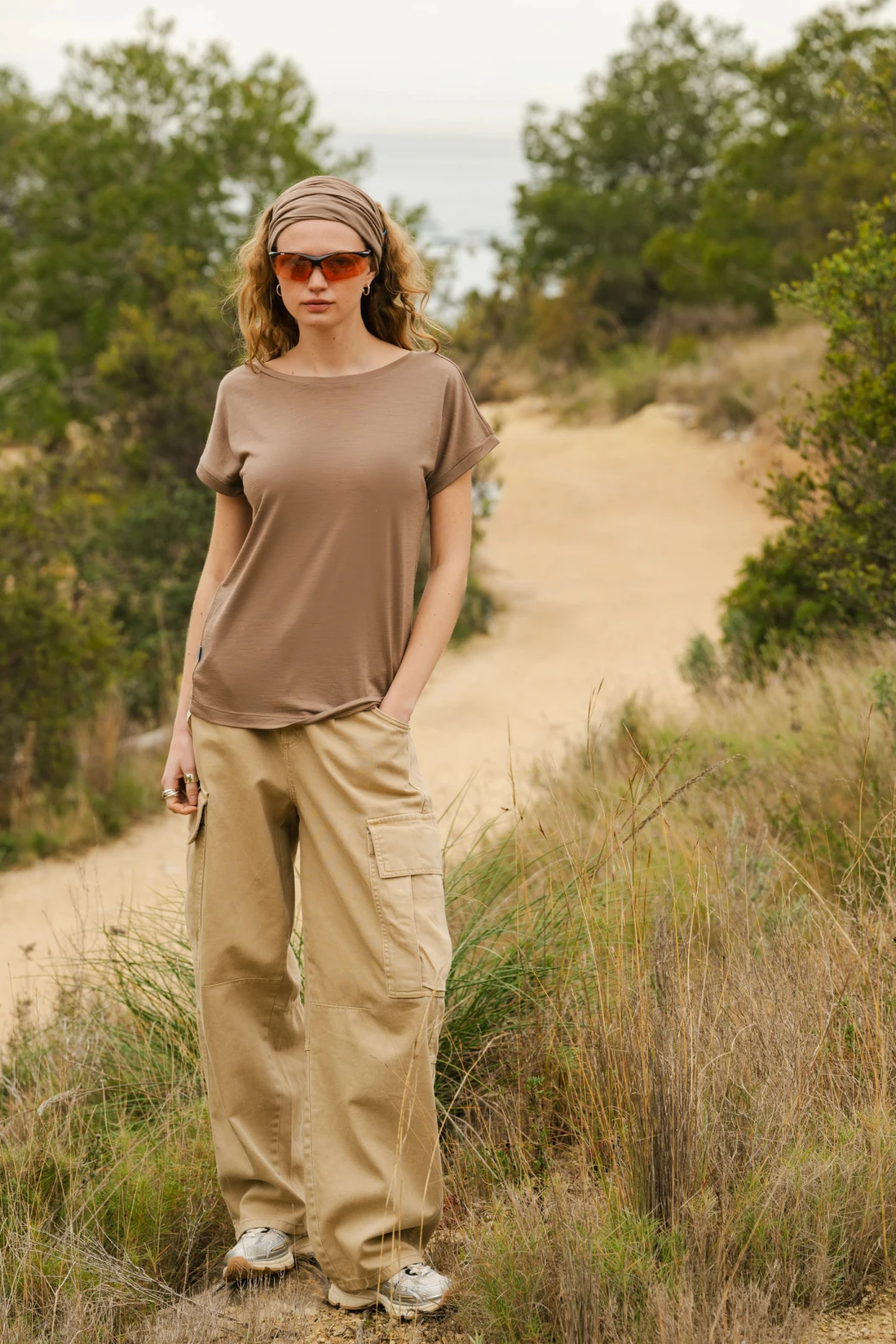 Osoba ubrana w szarą koszulkę, beżowe spodnie cargo, opaskę na głowę i pomarańczowe okulary przeciwsłoneczne stoi na zewnątrz na polnej ścieżce otoczonej wysoką trawą i zielonymi krzewami.