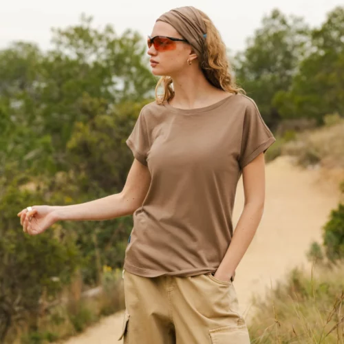 Kobieta w okularach przeciwsłonecznych, opasce na głowie w kolorze ciemnoszarym i brązowej koszulce z brązowymi spodniami stoi na polnej ścieżce otoczonej zielenią, patrząc w bok z jedną ręką w kieszeni.
