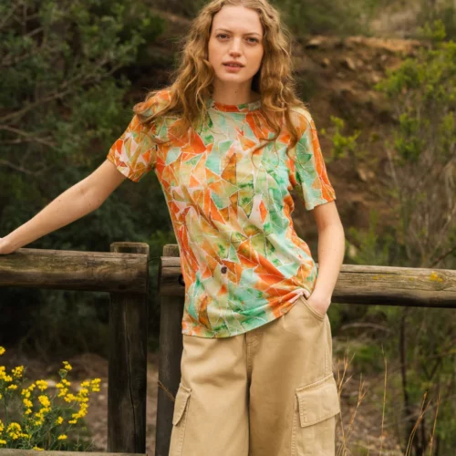 Kobieta z długimi falującymi włosami opiera się o drewniany płot na zewnątrz, ubrana w kolorową koszulkę z geometrycznymi wzorami i beżowe spodnie cargo. W tle widoczne są zielone drzewa i żółte kwiaty.