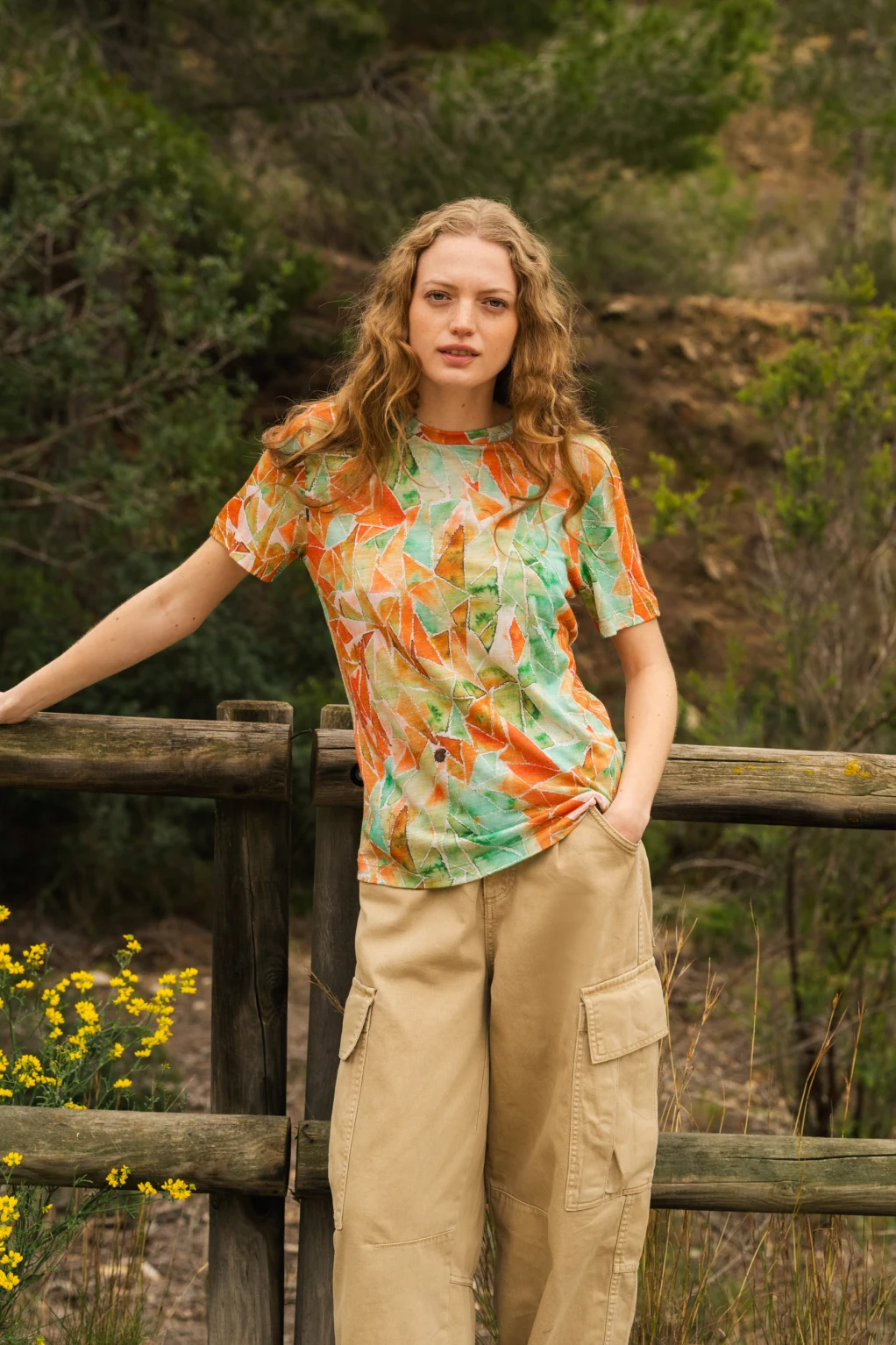 Kobieta z długimi falującymi włosami opiera się o drewniany płot na zewnątrz, ubrana w kolorową koszulkę z geometrycznymi wzorami i beżowe spodnie cargo. W tle widoczne są zielone drzewa i żółte kwiaty.