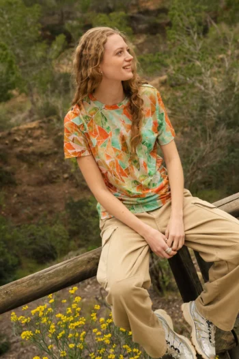 Osoba z długimi kręconymi włosami siedzi na drewnianym płocie na zewnątrz, uśmiechając się. Ma na sobie kolorową koszulkę z geometrycznym wzorem, beżowe spodnie i białe trampki. W tle widać żółte kwiaty i drzewa.