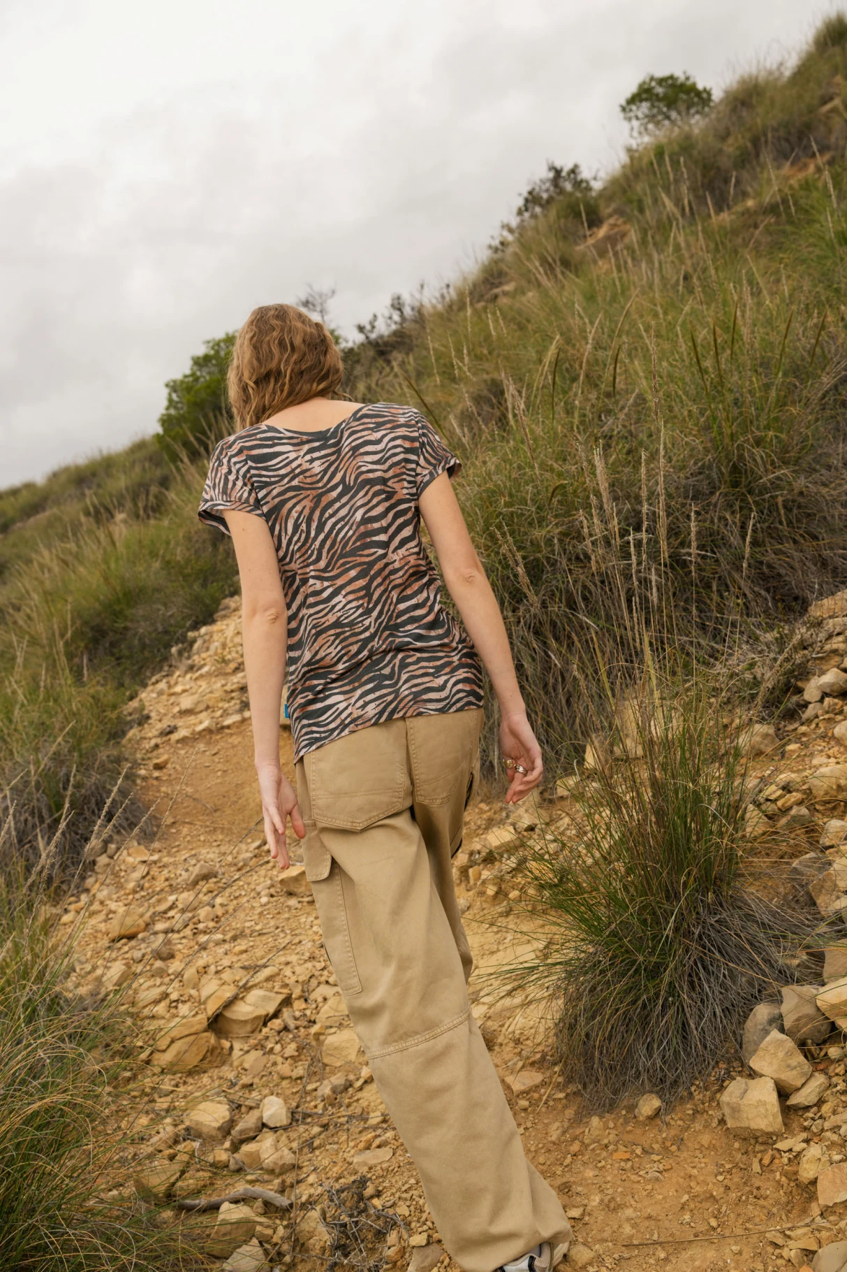 Osoba z krótkimi włosami, ubrana w koszulę z nadrukiem zebry i beżowe spodnie, idzie po skalistym, trawiastym wzgórzu w pochmurny dzień. Scena rozgrywa się na zewnątrz i jest widziana od tyłu.