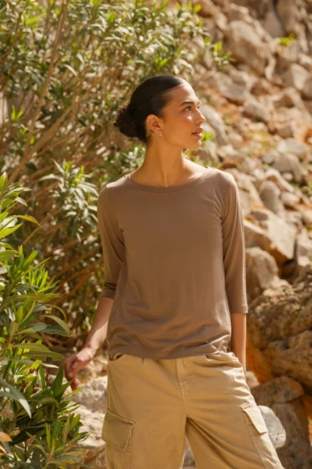 Kobieta z ciemnymi włosami związanymi z tyłu stoi na zewnątrz wśród zieleni i skalistego terenu, ubrana w Koszulkę z wełny merino z rękawem 3/4 mokka, patrząc spokojnie w lewo.