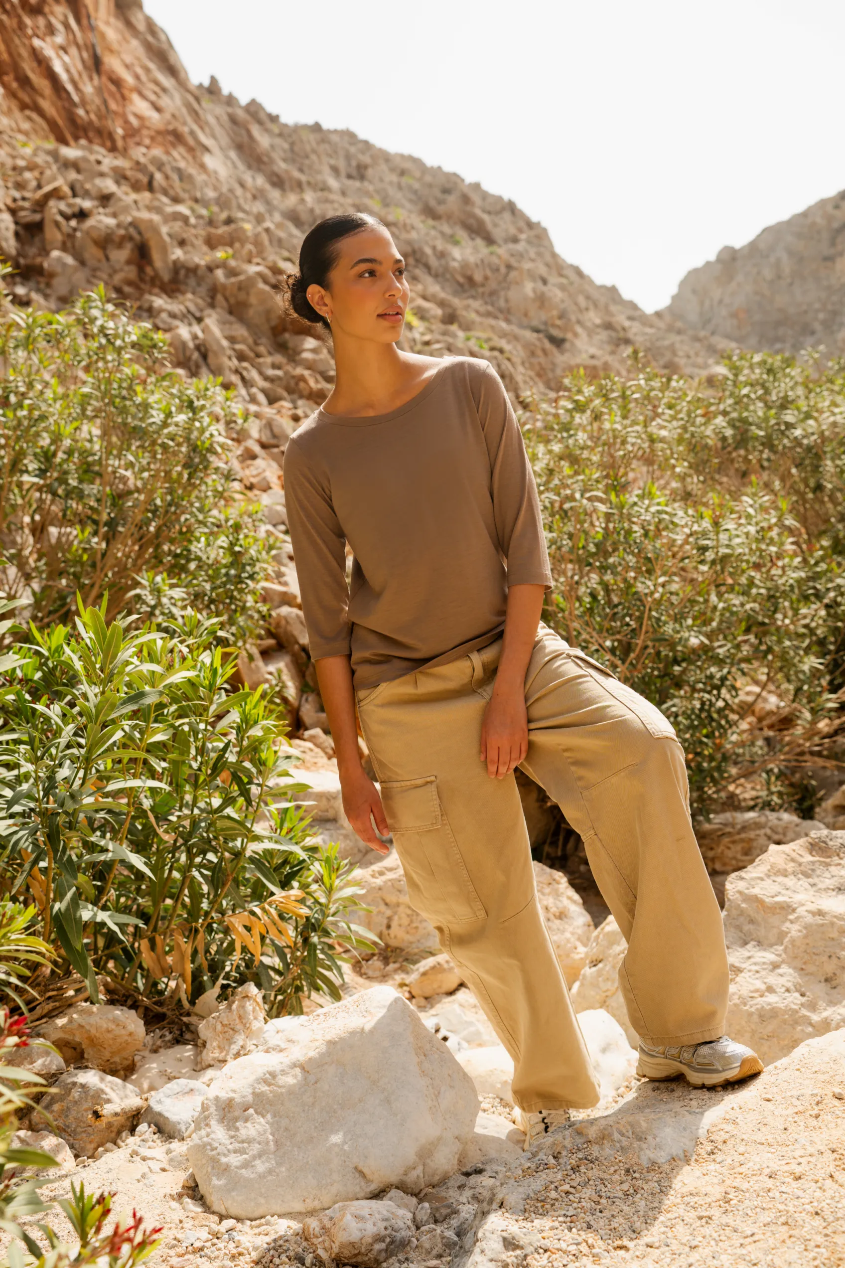 Osoba z ciemnymi włosami zaczesanymi do tyłu, ubrana w koszulkę z wełny merino z rękawem 3/4 mokka, stoi wśród skał i zielonych krzewów w oświetlonym słońcem, skalistym krajobrazie na zewnątrz.