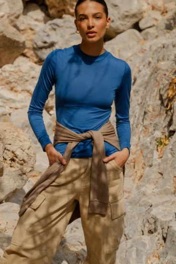 Na świeżym powietrzu wśród skał, osoba nosi Koszulkę z wełny merino z rękawem 3/4 mokka, z rękami w kieszeniach i neutralnym wyrazem twarzy.