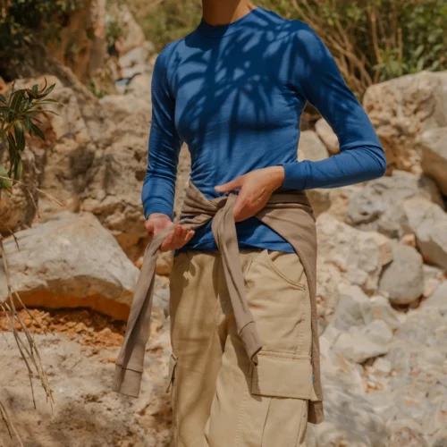 Ubrana w koszulkę z wełny merino z rękawem 3/4 mokka, osoba stoi na zewnątrz wśród skał i drzew, lekka kurtka przewiązana w talii, a cienie liści grają na jej twarzy i ciele.