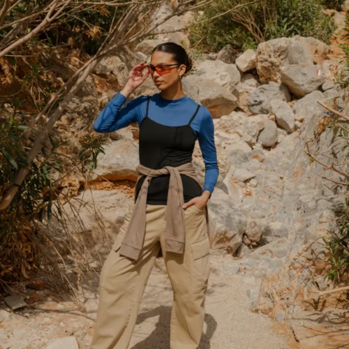 Kobieta stoi na kamienistym szlaku, ubrana w Koszulkę z wełny merino z rękawem 3/4 mokka pod czarnym tank, z beżowymi spodniami cargo, trampkami, okularami przeciwsłonecznymi w dłoni i kurtką przewiązaną w talii, patrząc w bok.