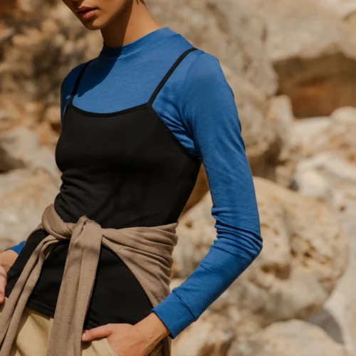 Kobieta stoi na zewnątrz na skalistym terenie, w okularach przeciwsłonecznych i koszulce z wełny merino z rękawem 3/4 mokka, pod czarnym tank topem ze spodniami khaki i beżowym swetrem zawiązanym w talii.