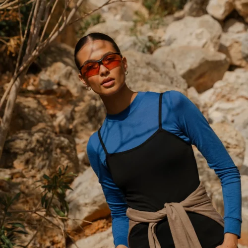 Kobieta w czerwonych okularach przeciwsłonecznych i koszulce z wełny merino z rękawem 3/4 mokka stoi na zewnątrz przed skalistym terenem i zielenią.