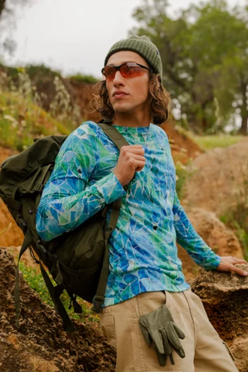 Mężczyzna na świeżym powietrzu stoi z dużym plecakiem, ma na sobie okulary przeciwsłoneczne i koszulkę z wełny merino z rękawem 3/4 mokka oraz zieloną czapkę. Zielone rękawiczki wystają z kieszeni jego spodni khaki, gdy opiera jedną rękę na kamieniu.