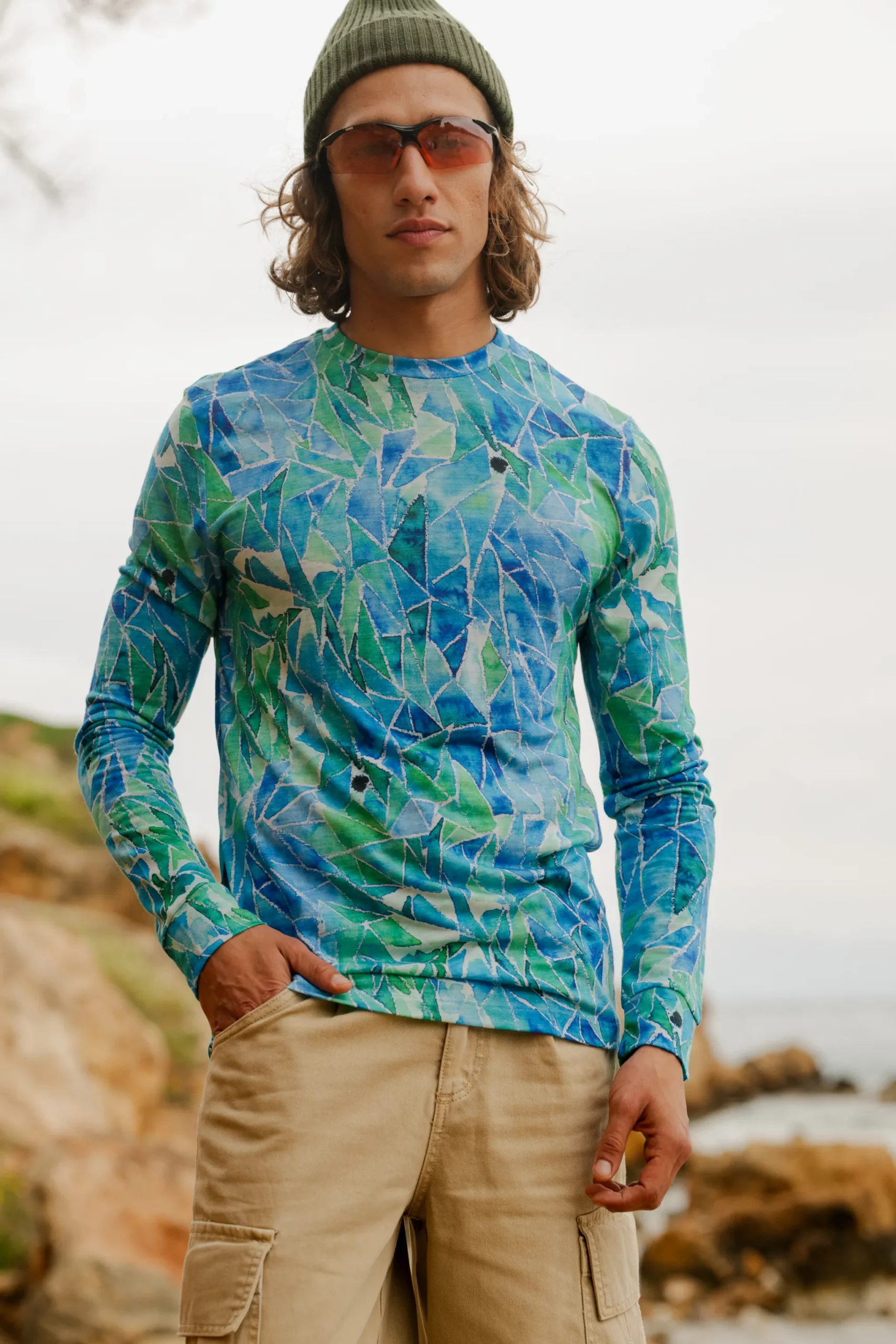 Osoba ubrana w okulary przeciwsłoneczne, zieloną czapkę, szorty khaki i koszulkę z wełny merino z rękawem 3/4 mokka stoi na zewnątrz w pobliżu skalistego terenu nad wodą.