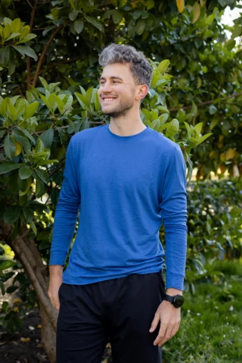 Kobieta ubrana w prostą koszulkę z wełny merynosów w kolorze mokka stoi uśmiechnięta na zewnątrz przed zielonymi krzewami liściastymi.