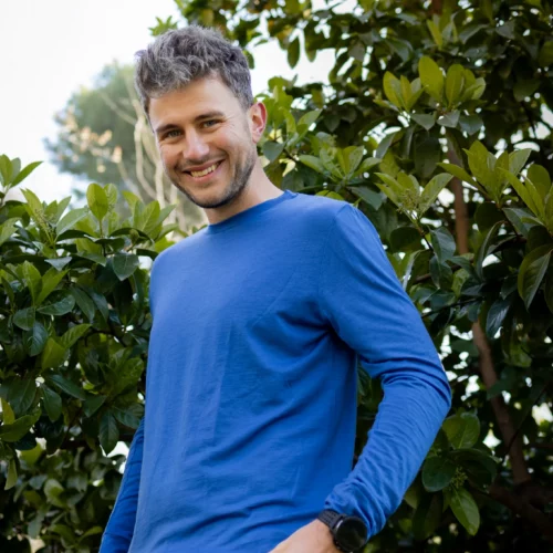 Mężczyzna z krótkimi siwymi włosami stoi na zewnątrz przy zielonych krzewach, ubrany w niebieską koszulę z długim rękawem i czarne spodnie obok Koszulki z wełny merino prosta damska mokka.