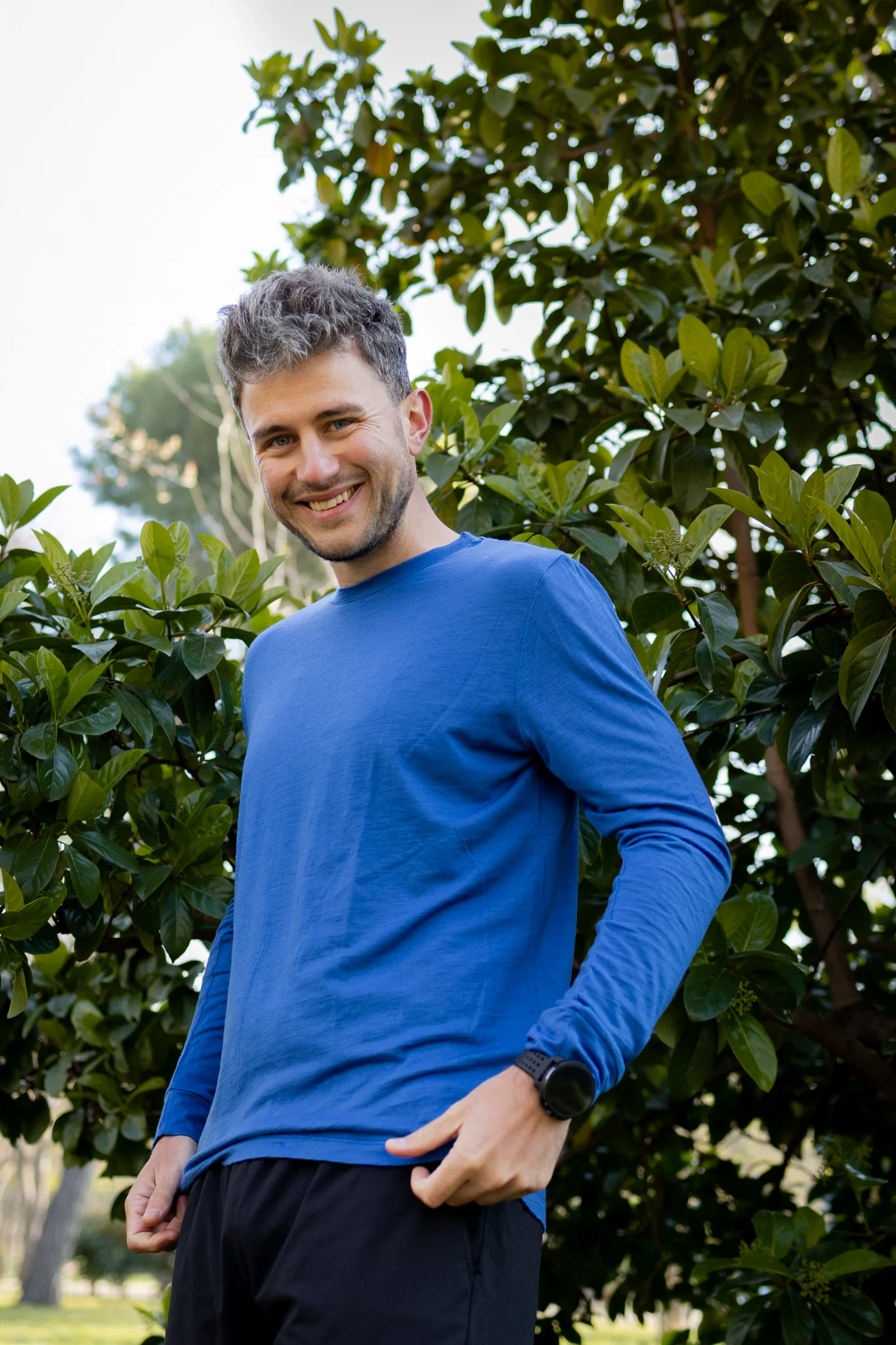 Mężczyzna z krótkimi siwymi włosami stoi na zewnątrz przy zielonych krzewach, ubrany w niebieską koszulę z długim rękawem i czarne spodnie obok Koszulki z wełny merino prosta damska mokka.