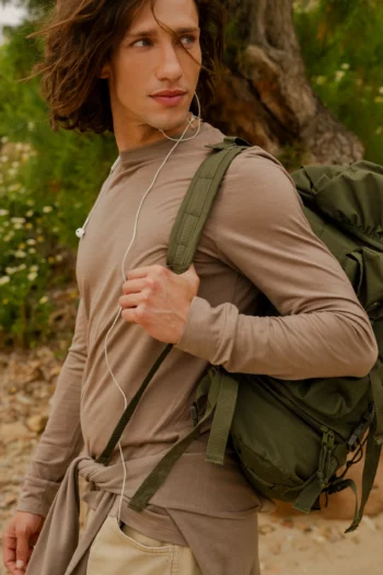 Młody mężczyzna z brązowymi włosami do ramion ma na sobie koszulkę z wełny merino z rękawem 3/4 mokka, spodnie khaki i zielony plecak. Stoi na zewnątrz na skalistej ścieżce z zielenią w tle, ze słuchawkami w uszach, patrząc w bok.