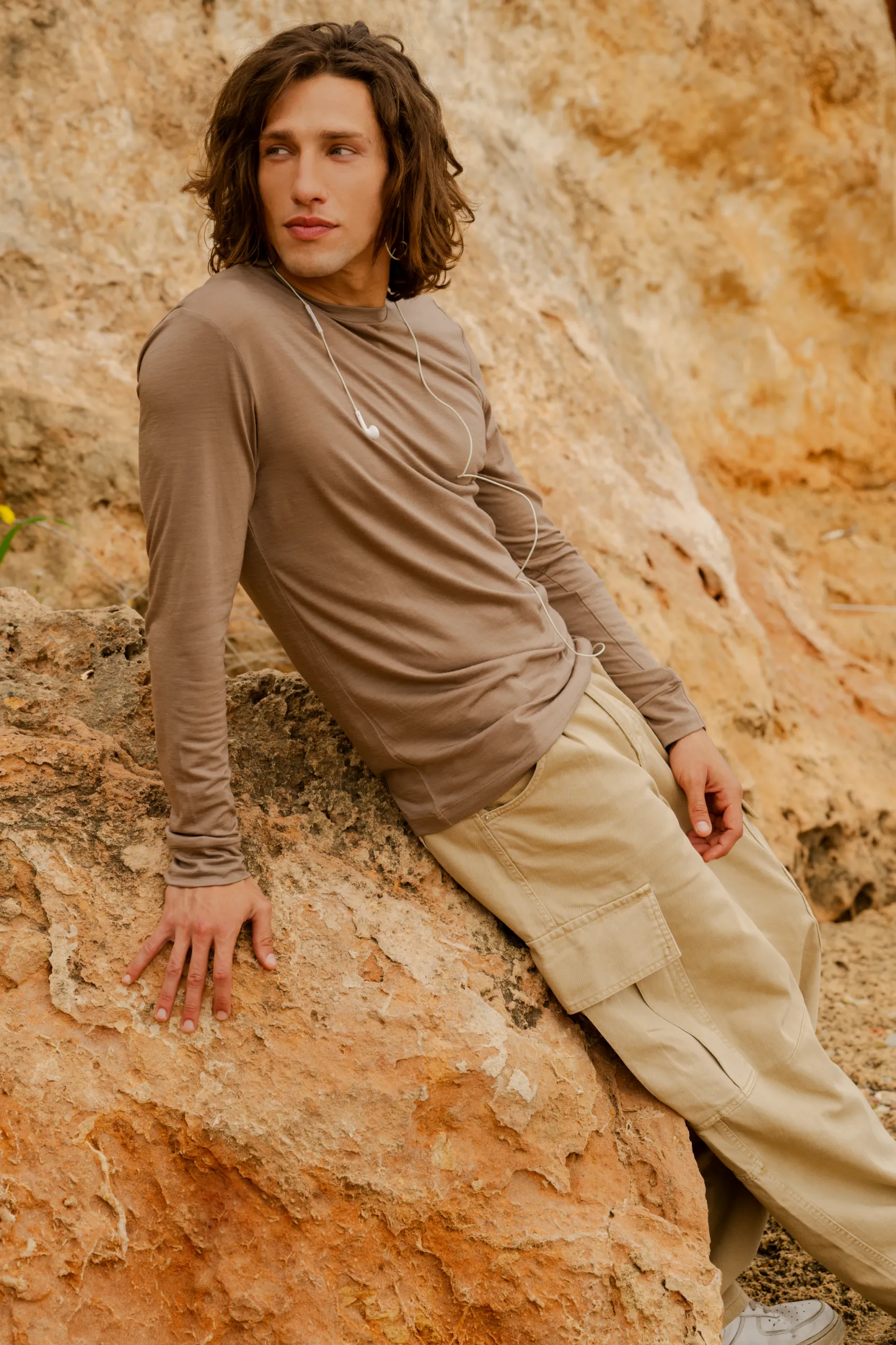 Młoda osoba z długimi brązowymi włosami, ubrana w koszulkę z wełny merynosów z rękawem 3/4 mokka, opiera się o skały na zewnątrz, patrząc w bok ze słuchawkami na szyi.