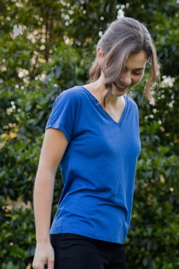 Kobieta z falowanymi włosami spiętymi w kucyk uśmiecha się, patrząc w dół, ubrana w koszulkę z wełny merino prosta damska mokka na zewnątrz z zielonymi liśćmi za nią.