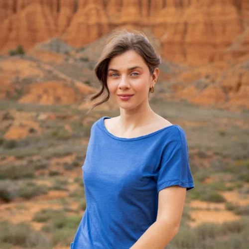 Kobieta o falowanych brązowych włosach nosi koszulkę z wełny merino prosta damska mokka, stojąc na zewnątrz wśród czerwonych formacji skalnych i rzadkiej roślinności, patrząc w kamerę z lekkim uśmiechem.